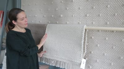 Taina Snellman-Langenskiöld står invid yllemattor i vitt och beige som hänger på en vägg. Hon tittar på mattorna och känner på en av dem.