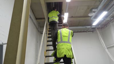 Två arbetsklädda män klättrar uppför en brant stege inne i en fabrik.