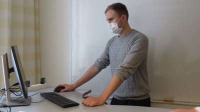 En man står vid en dator. Han har munskydd.