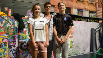 En äldre kvinnlig lärare står mellan två elever i 15-års åldern i ett aktivitetscenter med graffiti i bakgrunden.