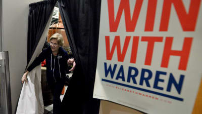 Kvinna går igenom gardiner till sitt kampanjmöte. Kvinnan är Elizabeth Warren. 