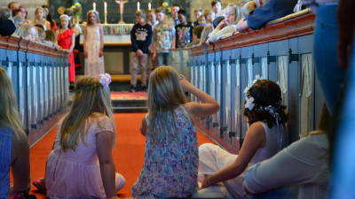 Några flickor sitter i altargången i en kyrka. Uppe vid altaret står uppklädda skolelever.