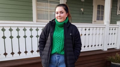 Ung kvinna i grön tröja och svart jacka utanför hus.