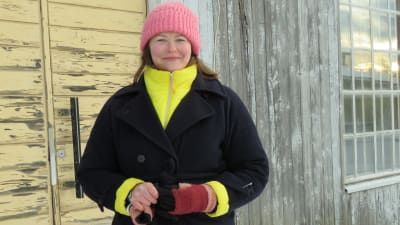 Nina Sointu i svart vinterrock och rosa mössa. Hon står ute. Vinter och bakom henne en trävägg i grått och gult. Färgen flagar.
