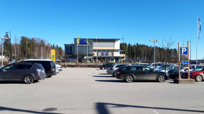 En parkeringsplats med bilar framför en affär. En person går förbi längre bort.