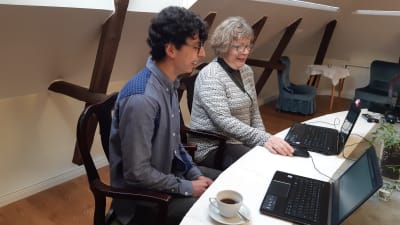 en ung mörkhårig man sitter på gammaldags stol bredvid en äldre medelålders kvinna. Framför sig på bordet har de två datorer, en grönväxt och en kaffekopp