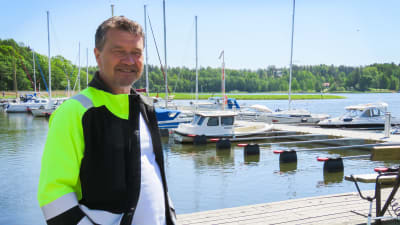 En man klädd i typiska arbetskläder i mörkblått och neon står framför en brygg med förtöjda båtar. Han ler och ser glad ut. Sommar och soligt.