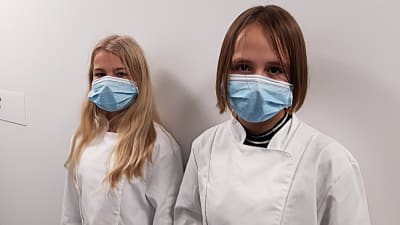 Två flickor i vita skyddsrockar och munskydd.