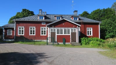 Ett rött ganska stort trähus. Sommar, sol, gröna träd och buskar. Sandplan framför huset.