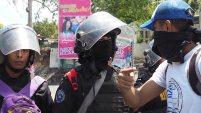 En ung demonstrant försöker förhandla med kravallpolis om att komma fram. I bakgrunden ett av otaliga enorma propagandaplakat med president Daniel Ortega och hustrun, vicepresident Rosario Murillo.