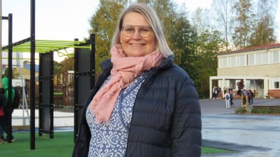 Porträttbild på Maria Lindberg, en kvinna med ljust, halvlångt hår, glasögon och svart, tunn dunjacka.  I bakgrunden en skolgård med elever. Höst.