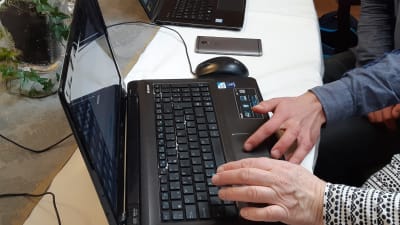 en äldre och en yngre hand ligger på tangenterna till en svart dator. En annan dator syns i bakgrunden och lite av en grönväxt