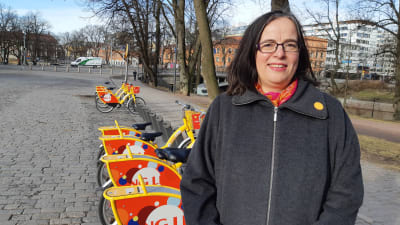medelålders mörkhårig kvinna i grå kappa står framför en rad gula stadscyklar på stentorg i Åbo