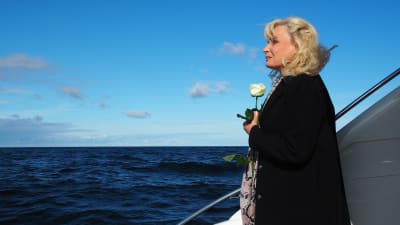 blond kvinna i mörkblå kappa står i båt på öppet hav med en vit ros i sina händer