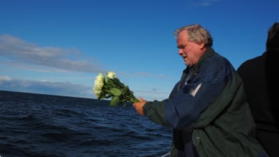 man står på båtdäck på öppet hav med bukett av vita rosor i händerna