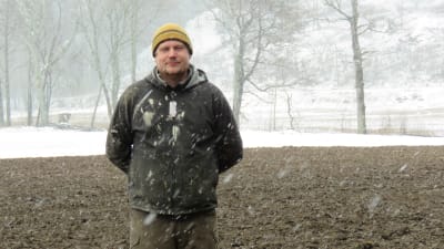 En man med gul mössa och grå jacka står på en åker som delvis är snötäck och delvis är bar. Det snöar ymnigt.