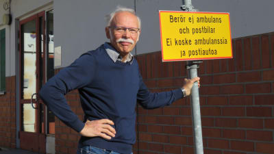 En man med grått hår, mustasch och blå tröja står utanför Ingå hälsocentrals ingång. Han står och håller i en trafikskylt som förbjuder trafik, men med tillägget "berör ej ambulans och postbil".