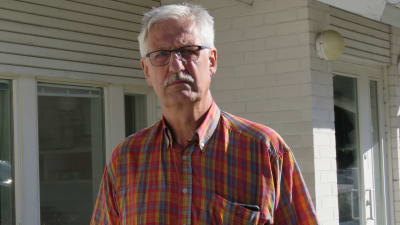 En allvarlig  man med grått hår, glasögon, mustasch och rödrutig skjorta framför ett vitt tegelhus