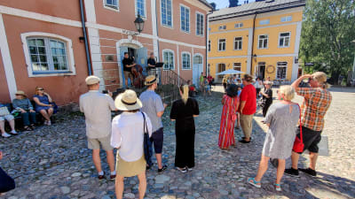 Folk står på ett torg och tittar upp på ett hus där en duo spelar musik på en trappa.
