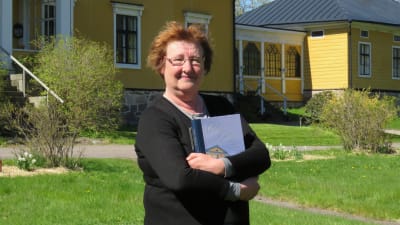 En kvinna med rött hår, glasögon och svart tröja håller om en bok. I bakgrunden ett stort gult hus. Vår, slutet av maj. Soligt.