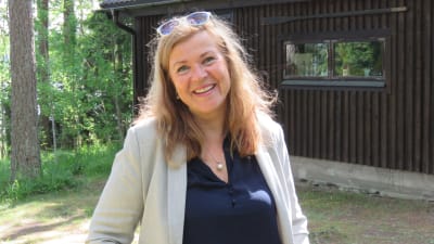 En kvinna med långt hår och solglasögon i hårfästet ler mot kameran. Hon har läppstift, blå skjorta och gråbeige linnekavaj. Hon ser glad ut. I bakgrunden en brun garagebyggnad eller egentligen ett hus.