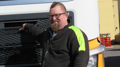 En man med kort och delvis snaggat hår, skägg och glasögon står framför en lastbilshytt. Han är klädd i arbetskläder i svart och neongult.