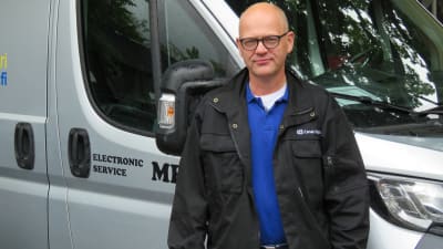 En man med glasögon,nästan utan hår står utanför en större paketbil.