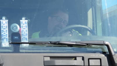 En man sitter inne i en lastbilshytt. Han pratar i telefon och ser ner på papper. Bilen står stilla.