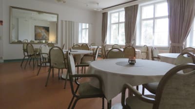 En matsal med stora fönster, stor spegel på ena väggen och flera mindre, runda bord med stolar runt borden.