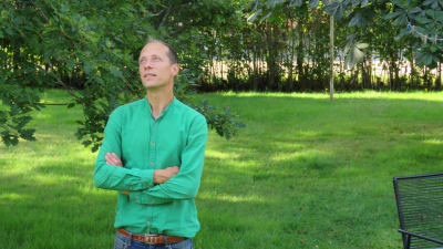En man i grön skjorta står i en grön och lummig trädgård. Han tittar uppåt, ser på ett hustak som inte syns på bild.
