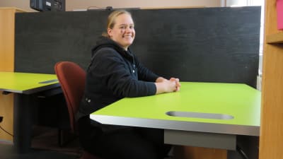 En kvinna sitter vid ett arbetsbord, på en kontorsstol i ett litet kontor tänkt för distansarbetare. Hon ler och ser in i kameran