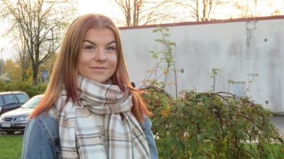 Portättbild på Linn Sjöberg, en tonåring med långt rödblont hår och jeansjacka. Soligt höstväder, lite dis. Kala träd och en grå cementbyggnad i bakgrunden.