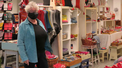 Äldre kvinna med munskydd står vid en hylla med massor av handarbeten och hantverk. Inne i en lokal.