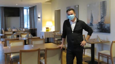 En man iklädd munskydd står i en tom, stor restaurang med dukade bord.
