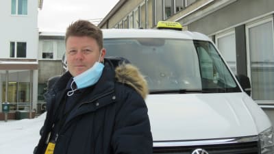 En man i blå jacka och munskydd hängande vid ena örat står utanför en stor vit taxi. Vinter, utanför Raseborgs sjukhus.