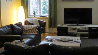 Ett vardagsrum med svarta lädersoffor och en tv. Plädar på stol och soffa och stearinljus på soffbordet. en tavla på väggen.