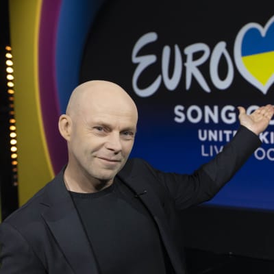 En skallig man i mörk kavaj står i en studio, ser in i kameran och pekar på logon för Eurovision Song Contest 2023.
