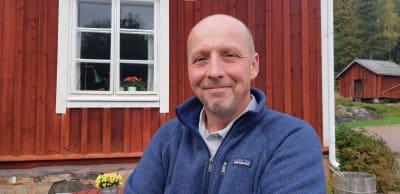 En glad man utan hår och med skägg i blå tröja står framför ett rött trähus med vita karmar och fönsterbågar.
