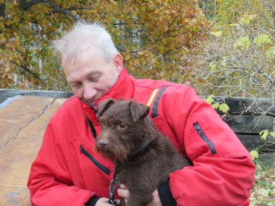 En man i röd rock håller en hund med brun lite längre päls i famnen. Höstgula lövträd i bakgrunden.