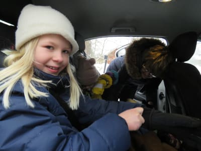 En blond flicka med vit mössa sitter i baksätet i en personbil. Hon ser in i kameran och ler. I baksätet finns också en yngre flicka som en äldre kvinna spänner fast säkerhetsbältet på.