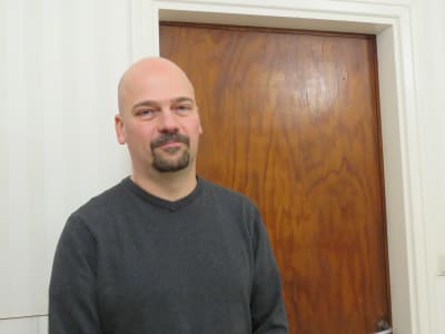 En man med rakat huvud och ett litet välansat skägg klädd i mörkgrå tröja står inne i en hall i ett hem. Han tittar in i kameran. Matias Kaihovirta.