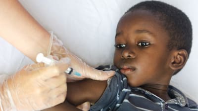 En liten pojke ligger på en säng och får vaccin i armen.