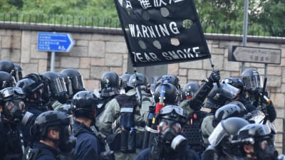 Poliser i svarta kläder och hjälmar håller i en skylt som varnar för tårgas