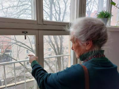 En äldre kvinna blickar och pekar ut genom ett högt ateljéfönster. Helsingfors breder ut sig mot Sörnäs framför henne.