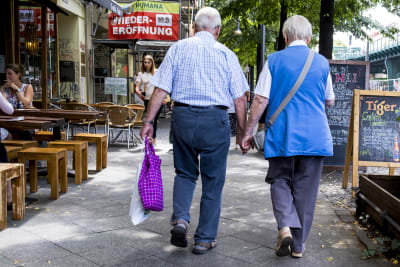 Ett äldre par  går hand i hand på en gatan med uteservering. 