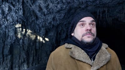 Sol strimmar in i grotta. Jan Jämsén med pälsrock, ser uppåt, uttryckslöst.