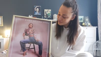 Artisten Kiara Nelson sitter på säng med en bild i ram föreställande hennes första singel. Hon har vita kläder. På bilden sitter hon på en stol.