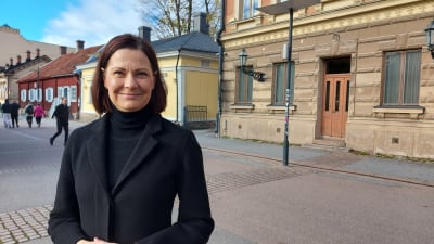 En leende svartklädd kvinna står på en gata framför ett stenhus.