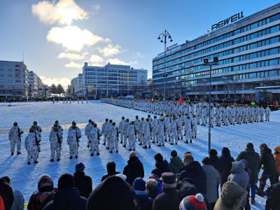 Solen lyser över tiotals beväringar som deltar i försvarsmaktens paradmönstring på Vasa torg. I förgrunden publik som samlats. 