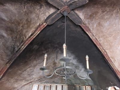 En elljuskrona hänger i en sakristia. Taket i sakristian är svart av sot efter en eldsvåda. Några inramade fotografier på väggen är också sotiga.